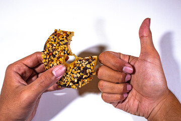 one donut with chocolate peanut topping, the tip has been bitten off, held in the left hand and the right hand with a thumbs up symbol, on a plain white backgorund - Powered by Adobe
