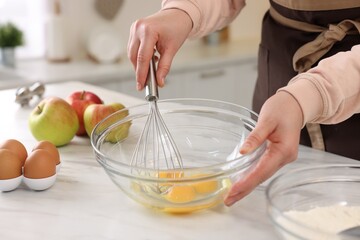 Woman whisking eggs in bowl at light marble table indoors, closeup