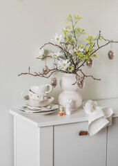 Ceramic white tableware, Easter decor, Easter bouquet on a white wooden chest of drawers in the living room. Holiday mood