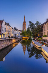 Fototapeta na wymiar Reflections on a canal in Bruges in the evening. Historic half-timbered houses and the Church of Our Lady in the center of the old town of the Belgian Hanseatic city. Boats and trees with footpath
