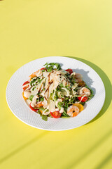 Fresh salad with greens, tomatoes, grilled shrimps on white plate. Top view on yellow background.