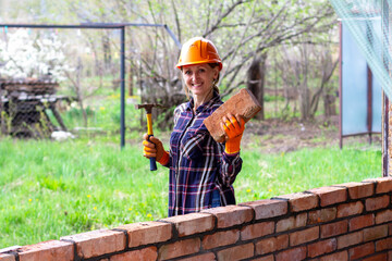 A young female bricklayer in an orange construction helmet with a construction hammer and a brick in her hands stands and smiles next to a brick wall