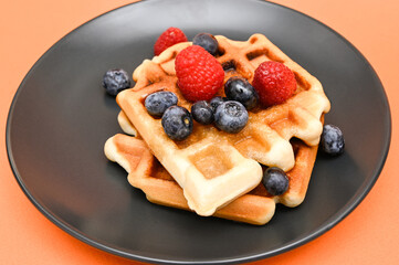 Belgian waffles and raspberries and blueberries on a black plate and on an orange background. Close-up.