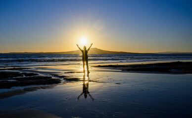 Woman raises arms towards the rising sun. Sunrise over the Rangitoto Island. Milford Beach....