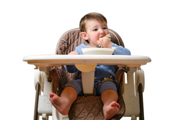 A funny child is eating a grated apple with his mouth full while sitting on a kitchen chair,...