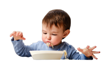 A funny child is eating a grated apple with mouth full while sitting on a kitchen chair, isolated on white background. Hungry baby boy shoves food in his mouth, humor. Kid aged one year four months