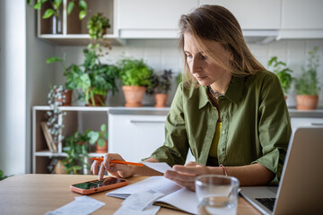 Pensive serious woman sitting at table, holding bills, receipts for utilities, credit payments,...