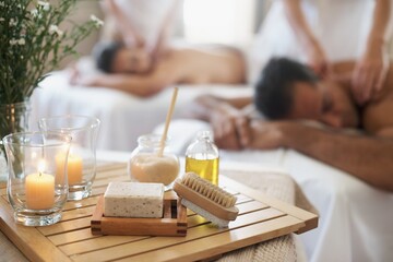 Candle, brush and couple in spa to relax on bed with luxury pamper treatment tools on table in...