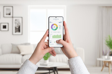 Woman holds smartphone with electricity usage app, monitoring and analyzing consumption for smart home. Living room background. Concept of energy efficiency - obrazy, fototapety, plakaty