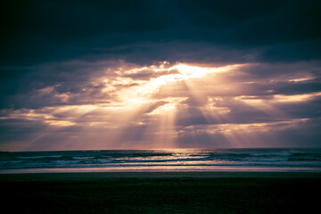 Sunset over Muriwai beach. Sun beams breaking through heavy clouds as waves splashing into wet sand. Auckland, New Zealand - obrazy, fototapety, plakaty