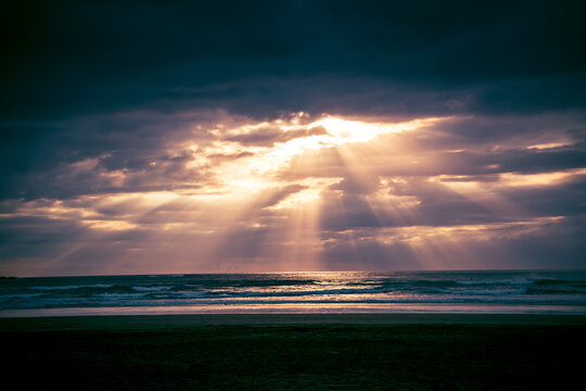 Sunset over Muriwai beach. Sun beams breaking through heavy clouds as waves splashing into wet sand. Auckland, New Zealand