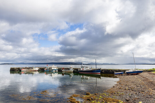 Gentle ripples intersect reflections at Broadford's shore, where boats rest by a stone pier, against a tapestry of Scottish isles under soft sunlight