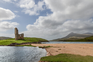 Ardvreck Castle stand on a green hillock by a calm lake, flanked by sandy beaches and distant mountains, under a vast sky of scattered clouds, epitomizing the scenic wilderness of Scotland