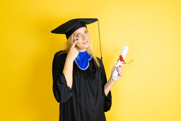 Beautiful blonde young woman wearing graduation cap and ceremony robe Pointing to the eye watching...