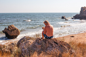 mature man sits on a rocky seashore with a cat in his hands. Traveling and tourism with pets