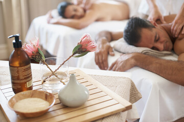 Hotel, flowers and couple in spa to relax on bed or break with luxury pamper treatment tools on table. Protea, facial oil or woman with man at resort or salon for natural healing benefits or massage - obrazy, fototapety, plakaty