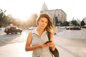Portrait of a young woman with curly hair against the backdrop of the city, she holds a phone in her hands. Sunny summer day.