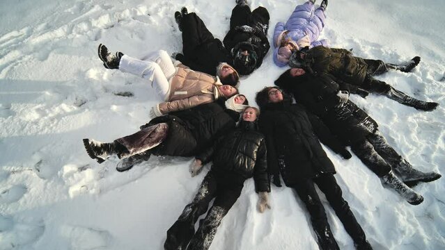 Friends lie in the snow on their backs in a circle shape.