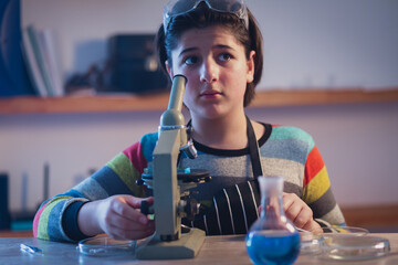 Cute young botanist in a home research laboratory wearing safety glasses sits near a microscope and looks up in surprise. - obrazy, fototapety, plakaty