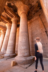 A blonde woman wearing a stylish hat examines the large columns at Karnak temple - Karnak temple in the heritage city of Luxor in Egypt. Giant row of columns with carved hieroglyph