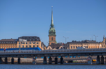 Naklejka premium Blue commuter train on a bridge passing the old town Gamla Stan and the church tower of Tyska kyrkan, a sunny winter day in Stockholm