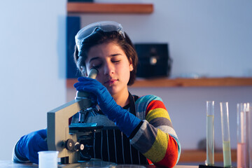 A modern Caucasian prodigy sits at a table in a home research laboratory and studies an object...