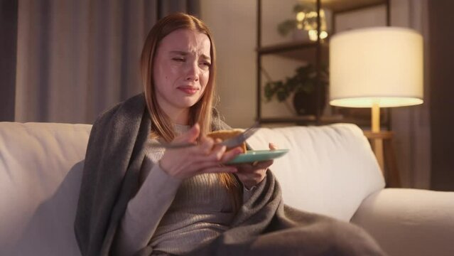 Portrait of sad young blond woman dealing with stress by eating food sitting on sofa late at home Upset female wrapped in blanket crying and eating cake at night indoors alone Mental heath problem