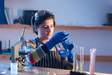 A modern Caucasian teenager sits at a table in a home research laboratory and intently studies the liquid in a flask. The child is wearing safety glasses, rubber gloves and an apron