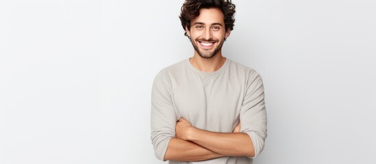 An attractive young, slim man is standing in front of a white wall with his arms crossed, looking satisfied and smiling. He appears to be relishing something good, as he rubs his hands together in