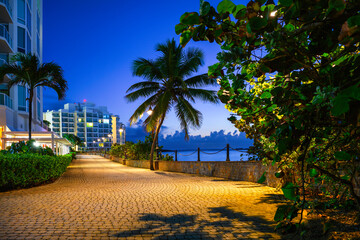 Fototapeta premium San Juan City Sunrise Landscape with tropical trees, buildings, and brick road along Pedestrian Shore Road around El Boquerón Bay and Balneario Condado Beach in Puerto Rico