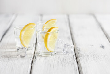 Shots of cold vodka with lemon and ice cubes on a white wooden table. Side view, selective focus, copy space. - 754284118