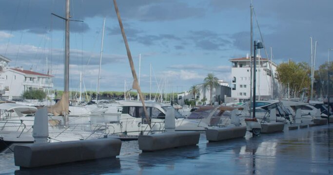 View on rainy port at small sea town in Europe. Boats and private yachts parked near the pier. Summer houses and property for rent. Wet asphalt and blue sky background. Luxury closed community. 