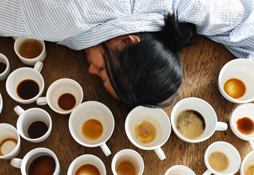 Exhausted, a tired female falls asleep at her working desk, surrounded by many empty coffee mugs, indicating a problem with caffeine addiction in female worker.