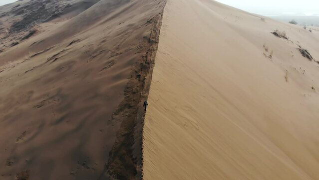 A man quickly climbs the crest of a huge dune. An aerial view taken from a drone. Singing barchan. Altyn-Emel National Park