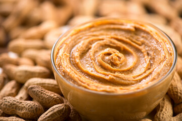 Peanut butter in a glass bowl over raw peanuts background. Creamy smooth peanut butter in glass bowl backdrop. Texture. Organic food. American cuisine