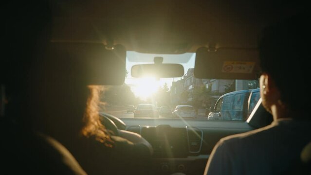 Rear view of a young couple driving a car in silence at sunrise or sunset. Sun blinds the female driver. Moving or traveling. Road trip with friends. Reaching destination. 