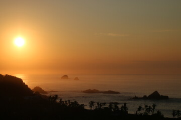 Puesta de sol en México, con la silueta de la costa, y el mar, con rocas y palmeras haciendo sombras .
