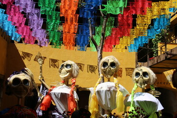 4 catrinas, esqueletos amarillos vestidos de mujeres, con trenzas, en un patio en Oaxaca, México, el día de muertos, con adornos de banderas de colores de papel picado y una pared amarilla. - obrazy, fototapety, plakaty
