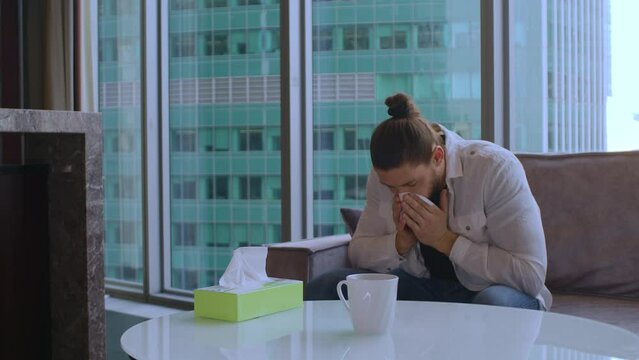 A man is sick, sitting on the couch at home and blowing his nose into a napkin. A man sick with the flu stayed at home and did not go out not to infect his colleagues. The man is sick and snotty.