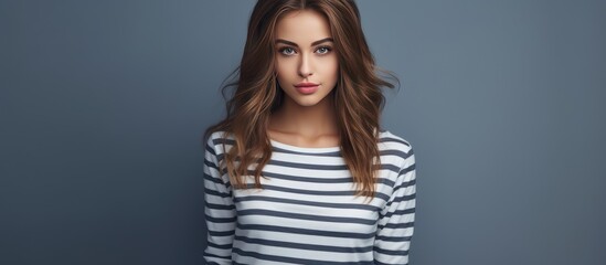 A beautiful young woman in a striped shirt poses confidently for a photograph on a grey background. Her posture is relaxed yet poised, exuding a sense of self-assurance and style.