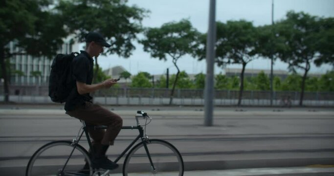 Stylish man rides minimalistic bicycle on the bike lane and checks smartphone for message or direction. Eco-friendly lifestyle. Young commuter reaching destination. Space for text. 