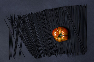 A succulent ripe Raf tomato on a black dyed spaghetti all on a smooth black surface - obrazy, fototapety, plakaty