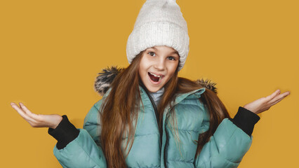 Little girl dressed in winter green down jacket and hat expresses surprise, shocked facial expression isolated on yellow background studio wall