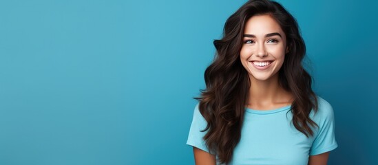 An attractive young woman with long dark hair is smiling and looking at the camera. She is standing in front of a blue background, wearing a blue shirt.