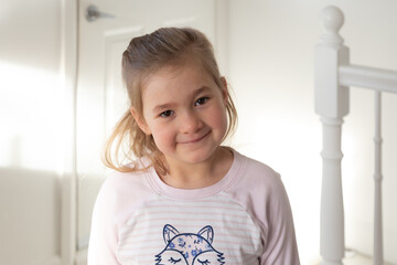 Cute happy smiling little five-year old girl standing in her pyjamas in room filled with morning...