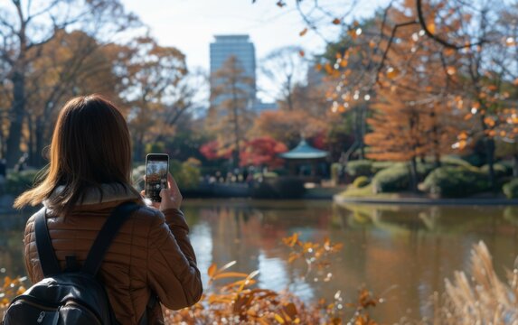 A woman is taking a picture of a pond with a cell phone. The pond is surrounded by trees and has a bridge in the background. The woman is wearing a brown jacket and a backpack