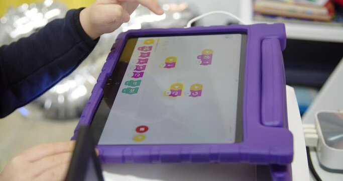Caucasian elementary school boy in glasses is concentrating assembling his own simple model of toy robot from plastic constructor on table. Details with electronic loop. Child sits in children room