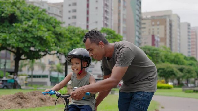 Happy latin father helping smiling boy to ride bike on cityu park. Happy latin child and young dad riding bike. Smiling daddy teaching son to ride a balance bicycle.
