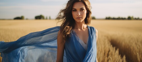 A woman wearing a blue dress is standing in a wheat field, with a blue shawl covering her shoulders. The field is golden and stretches out behind her under a clear sky.