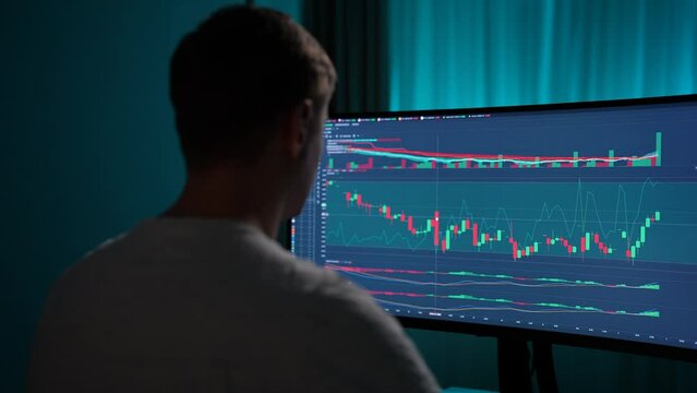 View from shoulder of concentrated crypto trader sitting in front of computers, making professional analysis of candlestick chart, creating strategy, looking at monitor in dark room, slow motion.